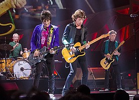 Rolling Stones performing on stage in Milwaukee, Wisconsin. From left: Charlie Watts on brown drum set, Ronie Wood wearing a purple jacket with black jeans playing a silver coloured guitar, Mick Jagger wearing black shirt and pants playing an orange/yellow guitar, Keith Richards with a green vest and black clothing playing an orange/yellow guitar (similar to Jagger's)