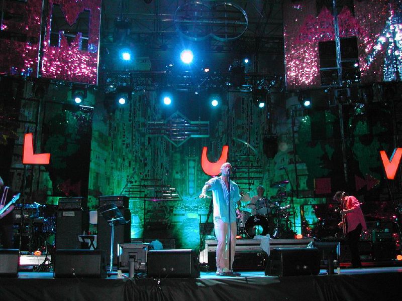 A blue-tinted photograph of musicians in front of an industrial background. From left to right: a long-haired male stands playing bass guitar, a middle-aged Caucasian male sings into a microphone, a middle-aged Caucasian male plays behind a black-and-silver drum set on a riser, and a guitar player at the edge of the photo.